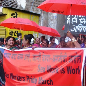 sex workers in India march through the street carrying red umbrellas saying "Sex work is work" and an NNSW banner